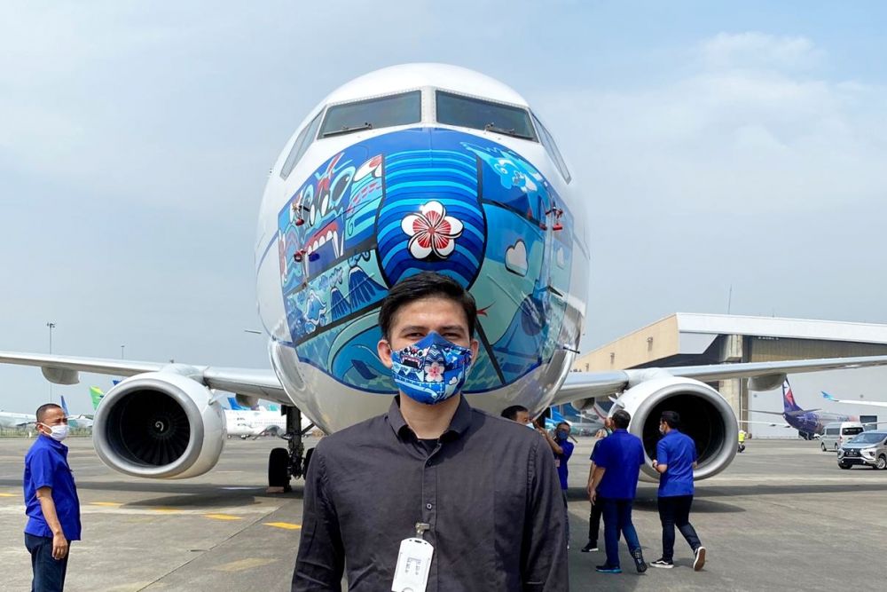 Jailani berdiri dengan latar belakang desain masker yang dibuatnya di pesawat Boeing 737-800NG Garuda Indonesia. FOTO: dok Jailani